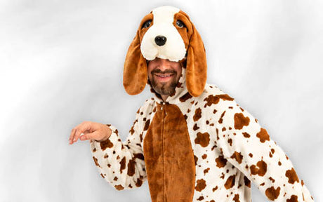Eine Person, die ein braun-weiß geflecktes Hundekostüm mit Schlappohren und einer großen Kapuze in Form eines Hundegesichts trägt und vor einem schlichten weißen Hintergrund steht.