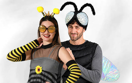 Zwei Erwachsene tragen Kostüme zum Thema Bienen, darunter Stirnbänder mit Fühlern, gestreifte Accessoires und große Insektenflügel, und posieren lächelnd vor einem schlichten weißen Hintergrund.