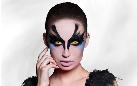 Person mit auffälligem Make-up in Schwarz und Blau, gelben Kontaktlinsen und einem Kostüm mit Federn, die mit einer Hand nahe am Gesicht vor einem hellen Hintergrund posiert.