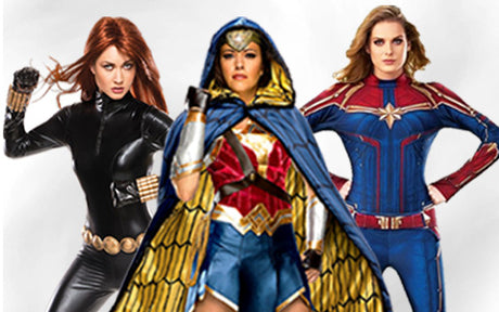 Drei Frauen in Superheldenkostümen posieren vor einem einfarbigen Hintergrund; eine in Schwarz, eine in Blau und Gold mit einem Umhang und eine in Rot, Blau und Gold.