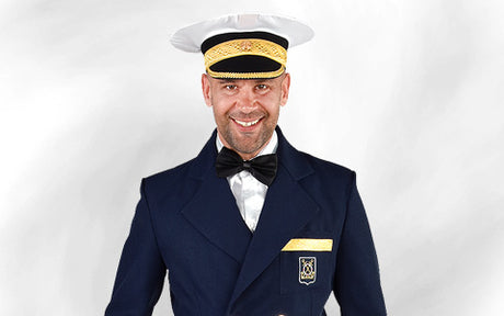 Ein Mann in einer marineblauen Uniformjacke, mit schwarzer Fliege und einem weiß-goldenen Hut, der vor einem schlichten, hellen Hintergrund steht und lächelt.