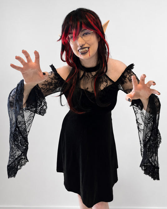 Eine Person in einem schwarzen Spitzenkleid posiert mit erhobenen Händen vor einem schlichten weißen Hintergrund; sie trägt Vampirzähne, spitze Ohren, eine Brille und eine Perücke mit roten Strähnchen.