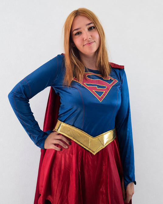 Eine Person in einem blau-roten Superheldenkostüm mit goldenem Gürtel und Umhang steht vor einem schlichten weißen Hintergrund und blickt in die Kamera.