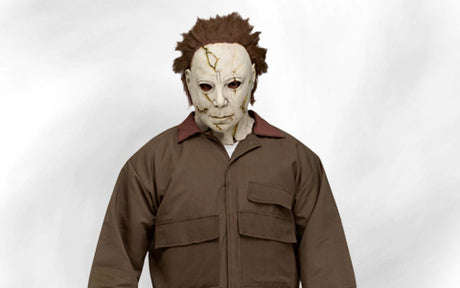 Eine Person mit einer weißen, rissigen Maske, zerzaustem braunem Haar und einem braunen Overall, die vor einem schlichten weißen Hintergrund steht.