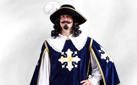 Eine Person in einem Musketierkostüm mit einem Federhut, einer lockigen Perücke, einem Schnurrbart und einem blauen Umhang mit goldenen Kreuzen, die vor einem schlichten weißen Hintergrund steht.
