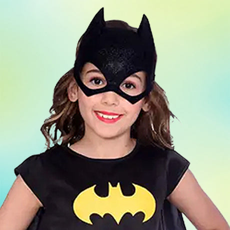 Ein Kind in einem schwarzen Batgirl-Kostüm mit Maske und einem gelben Fledermaus-Symbol auf der Brust lächelt vor einem hellblauen und grünen Hintergrund.