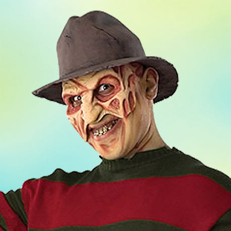 Eine Person, die einen Fedora-Hut und einen rot-grün gestreiften Pullover trägt, mit künstlichen Brandnarben und verfärbten Zähnen, die einer Figur aus einem Horrorfilm ähnelt, vor einem hellen Hintergrund.