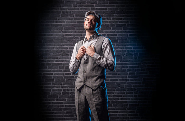 Ein Mann, der eine graue Weste, eine Hose, ein Hemd und eine Zeitungsjungenmütze trägt, steht vor einer dunklen Backsteinmauer und blickt mit den Händen auf der Weste nach oben.