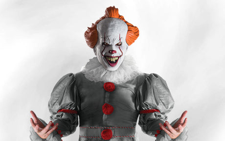 Eine Person in einem gruseligen Clownskostüm mit weißer Gesichtsbemalung, roter Nase, roten Haaren und spitzen Zähnen, die ein graues Outfit mit roten Knöpfen trägt und mit ausgebreiteten Armen vor einem weißen Hintergrund steht.