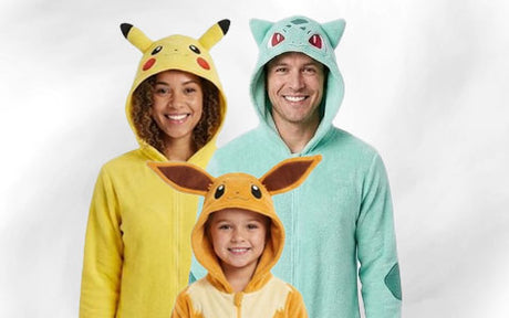 Drei Personen tragen Pokémon-Strampler: ein Erwachsener in einem Pikachu-Strampler, ein Erwachsener in einem Bulbasaur-Strampler und ein Kind in einem Eevee-Strampler; alle lächeln vor einem schlichten Hintergrund in die Kamera.