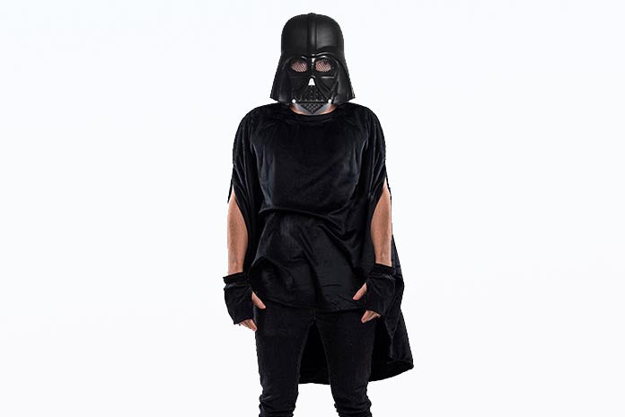 Eine Person, die vor einem weißen Hintergrund steht und einen schwarzen Umhang, Handschuhe sowie einen Helm trägt, der an Darth Vader aus „Star Wars“ erinnert.