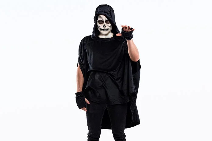 Eine Person in schwarzem Kostüm mit Totenkopf-Gesichtsbemalung und Kapuze, die vor einem weißen Hintergrund steht und eine Hand mit leicht gebeugten Fingern hebt.