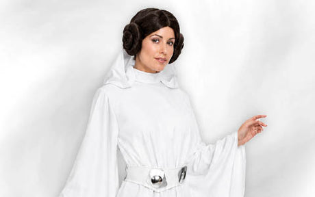 Eine Frau in einem weißen Gewand mit einem silbernen Gürtel und seitlichen Haarknoten, die vor einem schlichten weißen Hintergrund steht.