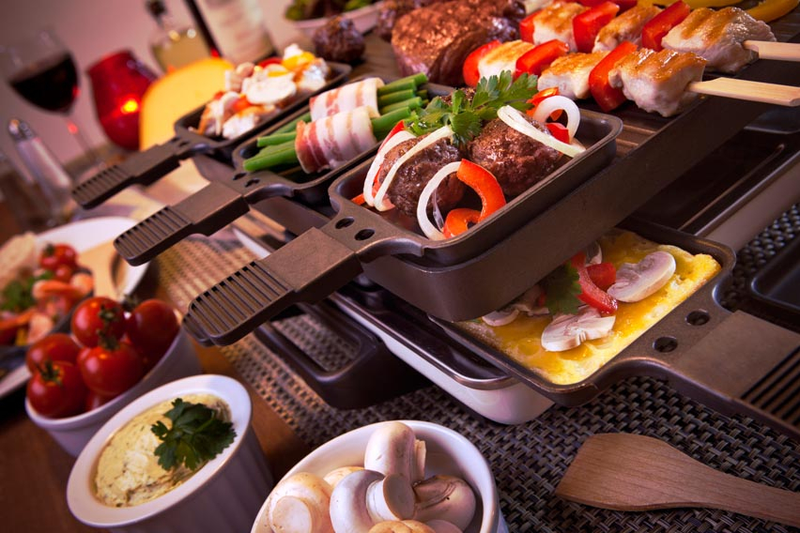 Ein Tischgrill, auf dem Fleisch- und Gemüsespieße liegen, um den herum auf dem Esstisch Käse, Pilze und andere frische Zutaten angeordnet sind.