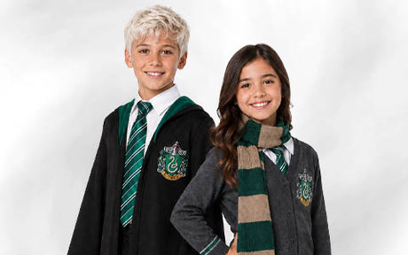 Zwei Kinder tragen Schuluniformen im Slytherin-Stil aus „Harry Potter“, darunter grüne und silberne Krawatten. Das eine trägt einen Umhang, das andere eine Strickjacke und einen Schal.