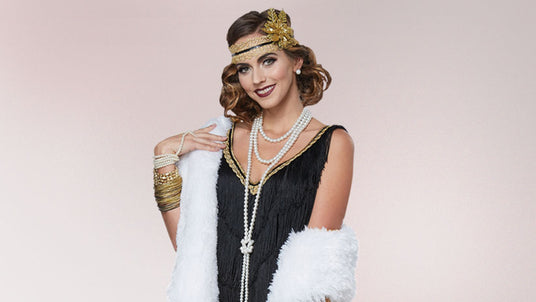 Eine Frau in einem Flapper-Kostüm aus den 1920er Jahren mit einem perlenbesetzten Stirnband, mehrreihigen Perlenketten, goldenen Armreifen und einem weißen Schal vor einem hellen Hintergrund.
