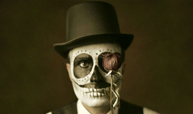 Eine Person mit Totenkopf-Gesichtsbemalung und Zylinder hält vor einem Auge eine verwelkte Rose in der Hand, vor einem dunklen Hintergrund.