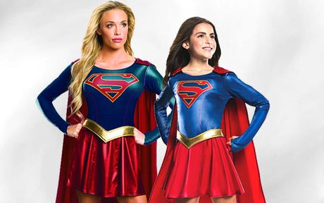 Zwei Personen in roten und blauen Supergirl-Kostümen mit Umhängen stehen nebeneinander vor einem hellen Hintergrund.