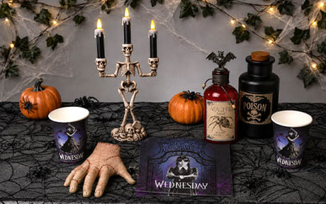 Tischdekoration zum Thema Halloween mit Skelett-Kandelabern, Kürbissen, künstlichen Zaubertränken, Spinnennetz-Tischdecke, Bechern, einer künstlichen, genähten Hand und einer Karte mit einer Gothic-Figur namens Wednesday.