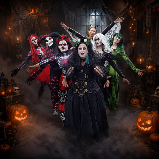 Sechs Personen in aufwendigen Halloween-Kostümen posieren mit ausgestreckten Armen in einem dunklen, nebligen Raum, der mit Kürbissen, Totenköpfen, Kerzen und Spinnweben geschmückt ist.