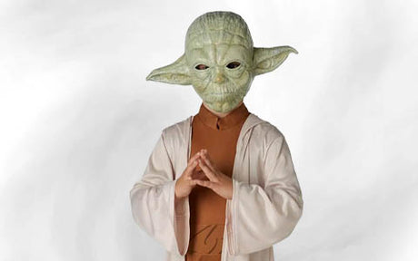 Eine Person, die eine Yoda-Maske und ein Yoda-Kostüm trägt, steht mit gefalteten Händen vor einem schlichten weißen Hintergrund.