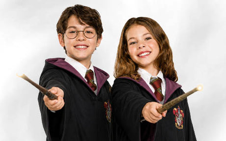 Zwei Kinder in Hogwarts-Roben, die Krawatten tragen und Zauberstäbe mit leuchtenden Spitzen in den Händen halten, lächeln vor einem schlichten weißen Hintergrund in die Kamera.