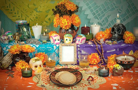 Ein farbenfroher Altar zum „Día de los Muertos“ mit Kerzen, Ringelblumen, Zuckerschädeln, einem gerahmten Foto, Essen, Getränken und „Papel Picado“-Dekorationen.