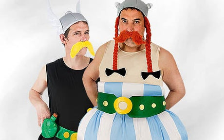 Zwei Erwachsene tragen Wikinger-Kostüme im Cartoon-Stil mit Helmen, bunten Schnurrbärten und übertriebenen Outfits; einer hat einen gelben Schnurrbart, der andere rote Zöpfe und ein gepolstertes, gestreiftes Outfit.