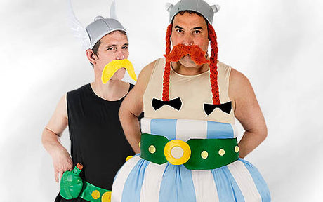 Zwei Erwachsene tragen karikaturhafte Wikinger-Kostüme mit Helmen, breiten Gürteln und falschen Schnurrbärten. Einer trägt ein gepolstertes Outfit mit blauen und weißen Streifen. Beide stehen vor einem schlichten, hellen Hintergrund.