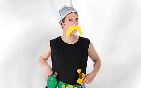 Eine Person, die einen Wikingerhelm mit weißen Flügeln, einen gelben falschen Schnurrbart, ein schwarzes, ärmelloses T-Shirt und ein Spielzeugschwert an einem grün-gelben Gürtel trägt.