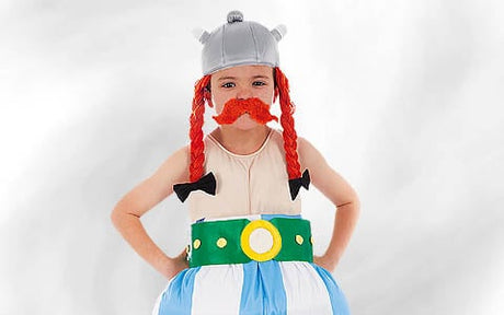 Ein Kind in einem Kostüm im Wikinger-Stil mit einem gehörnten Helm, einer roten, geflochtenen Perücke, einem falschen Schnurrbart und einem gepolsterten, gestreiften Outfit mit einem grünen Gürtel, das vor einem schlichten, hellen Hintergrund steht.
