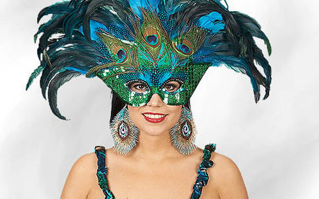 Eine Frau mit einer grün-blauen Maske aus Pfauenfedern, passenden Ohrringen und einem passenden Kleid, die vor einem schlichten weißen Hintergrund lächelt.