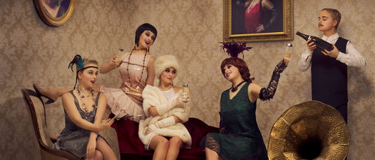 Fünf Personen in Outfits im Stil der 1920er Jahre posieren in einem Raum im Retro-Stil mit Champagnergläsern, einem Grammophon und einem gerahmten Porträt an der Wand.