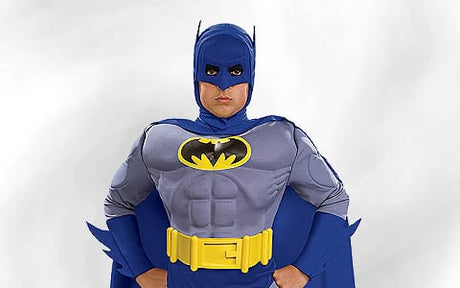 Eine Person in einem klassischen Batman-Kostüm mit gelbem Gürtel, blauem Umhang und Fledermaus-Symbol auf der Brust, die vor einem schlichten, hellen Hintergrund steht.