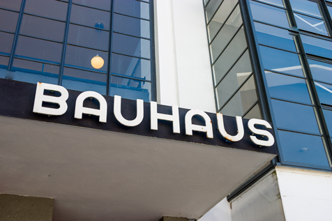 Bauhaus-Schild an der Fassade eines modernistischen Gebäudes aus Glas und weißem Beton mit geometrischen Fenstermustern.