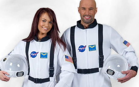 Zwei Personen in weißen Astronautenanzügen mit Aufnähern im NASA-Stil, die Helme in der Hand halten und in die Kamera lächeln. Auf jedem Ärmel befindet sich ein Aufnäher mit der amerikanischen Flagge.