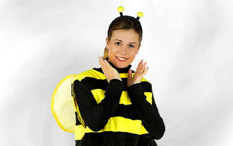 Eine Person in einem gelb-schwarzen Bienenkostüm mit Fühlern und Flügeln, die lächelt und vor einem schlichten, hellen Hintergrund posiert, wobei sie die Hände nahe am Gesicht hält.