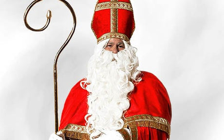 Eine als Nikolaus verkleidete Person trägt ein rotes Gewand und eine hohe rote Mütze und hält einen goldenen Stab vor einem schlichten weißen Hintergrund in der Hand.