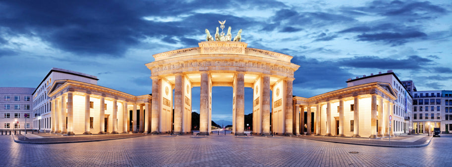 Das Brandenburger Tor in Berlin, Deutschland, bei Einbruch der Dämmerung beleuchtet, mit bewölktem Himmel im Hintergrund und einem leeren Kopfsteinpflasterplatz davor.