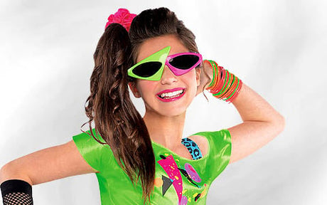 Eine lächelnde Person in einem leuchtend grünen Outfit, mit einer neonpinken und grünen eckigen Sonnenbrille, einem pinken Haargummi, bunten Armbändern und einem seitlichen Pferdeschwanz, die vor einem schlichten weißen Hintergrund posiert.