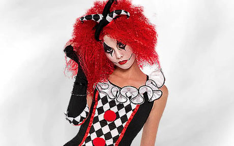 Eine Person in einem schwarz-weiß-roten Harlekin-Kostüm mit Rüschenkragen, auffälligem Make-up und einer leuchtend roten Lockenperücke vor einem schlichten weißen Hintergrund.