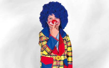 Eine Person in einem bunten, karierten Clownskostüm mit einer blauen Afro-Perücke und einer roten Nase, die lächelt und ihre Nase berührt, vor einem schlichten weißen Hintergrund.