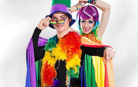Zwei Personen tragen farbenfrohe Regenbogen-Outfits, Umhänge und Accessoires, posieren gemeinsam und lächeln; die eine trägt einen lila Hut und eine Sonnenbrille, die andere eine knallbunte Perücke.