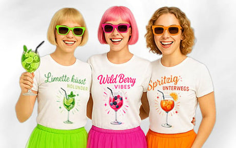 Drei Frauen in bunten Röcken und Sonnenbrillen lächeln; jede trägt ein weißes T-Shirt mit einem anderen Motiv eines Fruchtgetränks und einem deutschen Spruch und steht vor einem schlichten weißen Hintergrund.