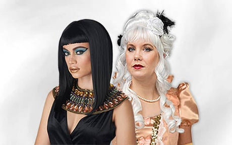 Zwei kostümierte Frauen: Die eine ist als Kleopatra verkleidet, mit schwarzer Perücke und auffälligem Make-up, die andere trägt ein Kleid im Stil des 18. Jahrhunderts mit weißer Perücke, Perlen und einem dekorativen Haarschmuck; beide stehen nebeneinander.