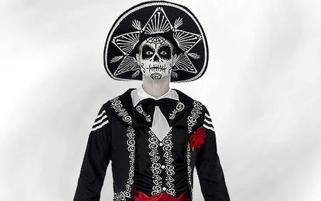 Eine Person in einem Kostüm im Mariachi-Stil mit Gesichtsbemalung im Stil des „Tages der Toten“, einem großen bestickten Hut und einer roten Schärpe, die vor einem schlichten hellen Hintergrund steht.