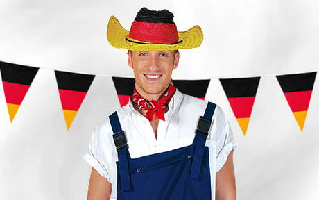 Ein Mann in blauer Latzhose, weißem Hemd und rotem Bandana, der einen Hut in den Farben der deutschen Flagge trägt, steht vor einer Girlande aus deutschen Flaggen.