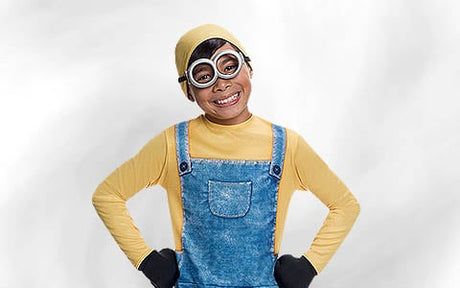 Ein als Minion verkleidetes Kind, das ein gelbes Shirt und eine gelbe Mütze, eine blaue Latzhose, schwarze Handschuhe und eine runde Schutzbrille trägt, steht mit den Händen in die Hüften gestemmt vor einem schlichten, hellen Hintergrund und lächelt.