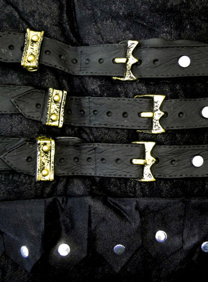 Drei schwarze Lederriemen mit verzierten goldenen Schnallen liegen waagerecht auf einer schwarzen, strukturierten Oberfläche über einer Reihe silberner Nieten.