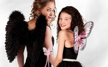 Zwei Mädchen in schwarzen Outfits stehen nebeneinander; das eine hat schwarze Engelsflügel und lockiges Haar, das andere hat Schmetterlingsflügel und einen Sternenstab. Beide blicken über die Schulter zurück und lächeln.